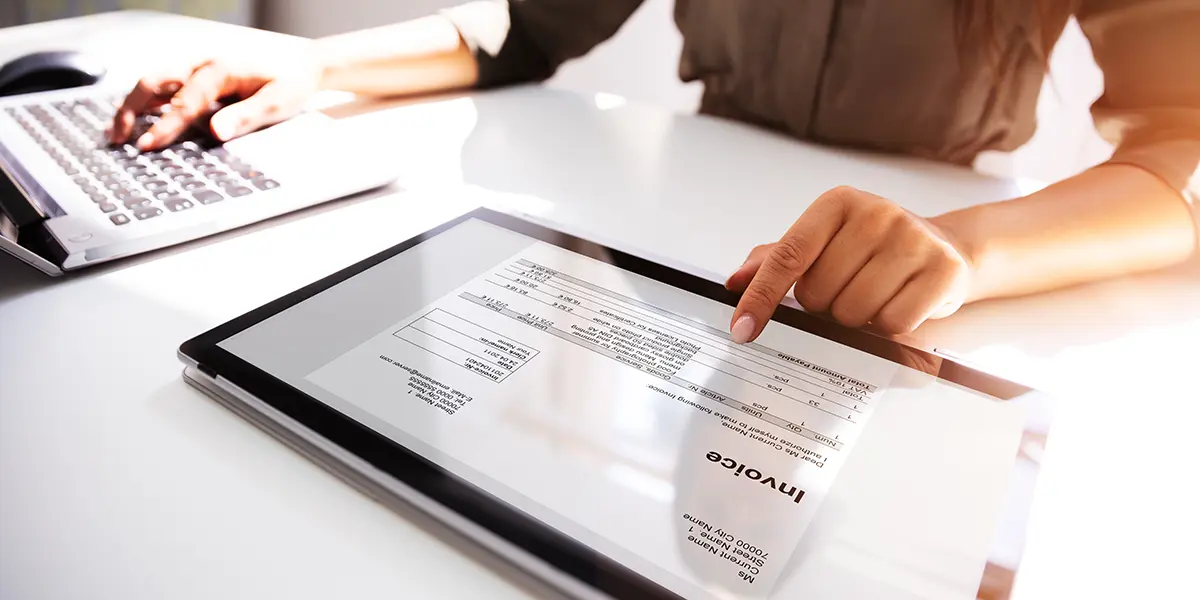 Woman looking at invoice on her tablet and preparing to pay for it using laptop